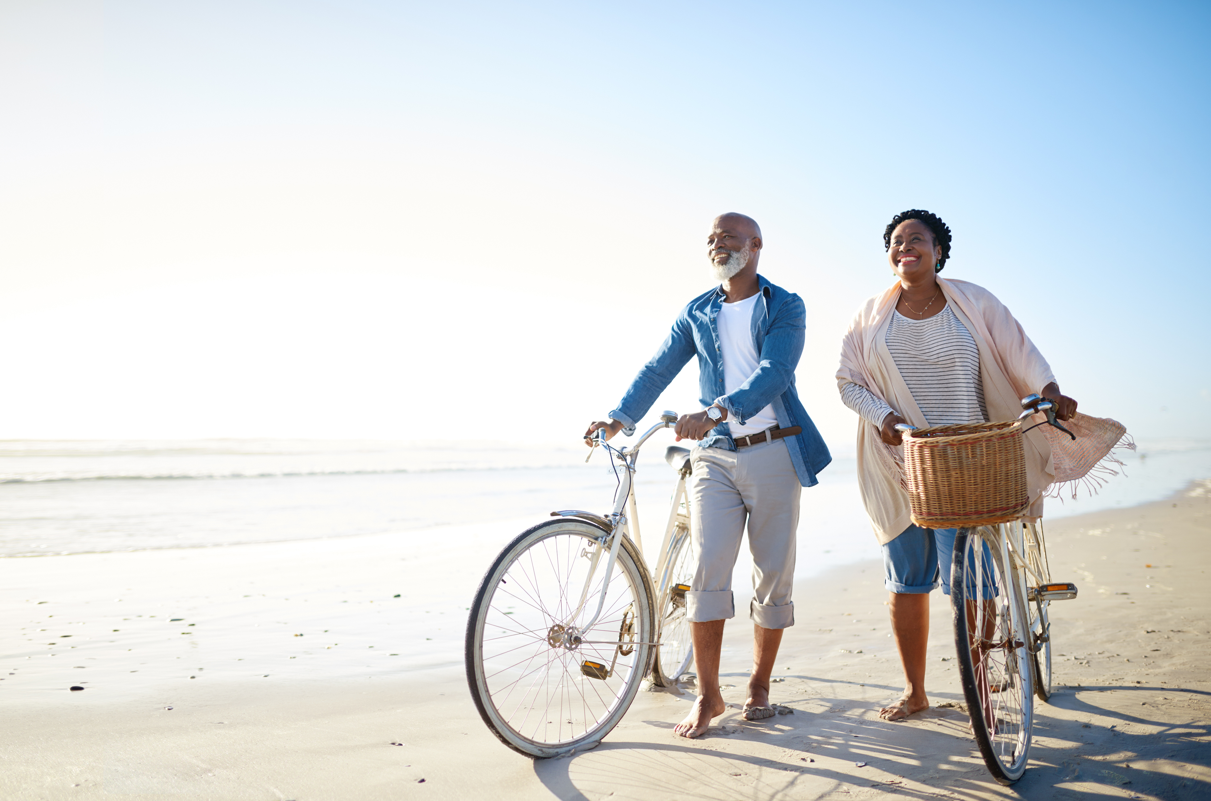 elderly married couple walking on beach with bicycles and smiling