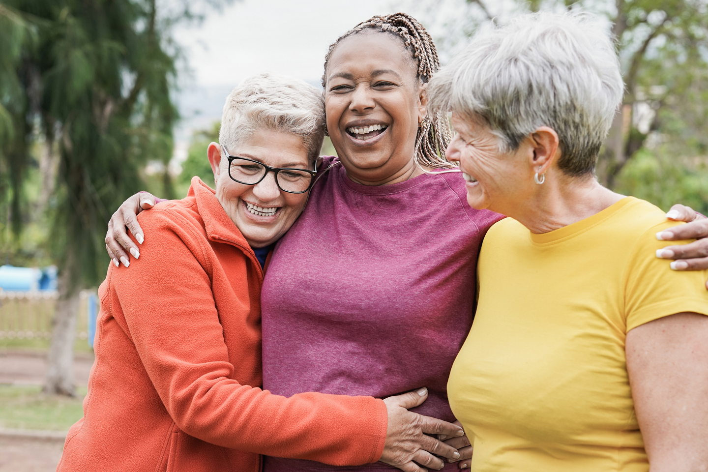 a trio of elderly women go walking on park trail and hug, smiling